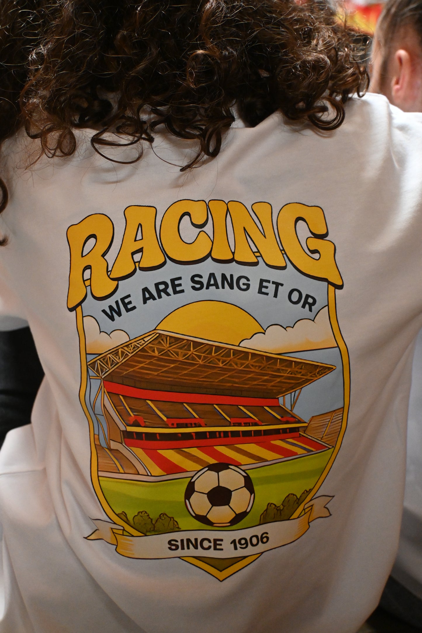 Dos d’un t-shirt blanc Enfant du Racing avec grand visuel coloré représentant le stade Bollaert, le mot “Racing”, le slogan “We are Sang et Or” et la mention “Since 1906”.