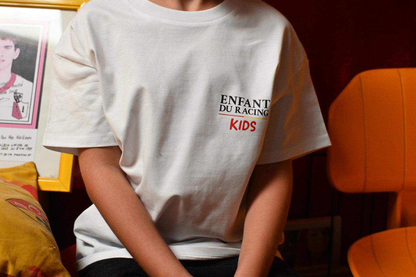 Enfant portant un t-shirt blanc Enfant du Racing Kids avec logo brodé côté cœur, assis dans un décor rétro aux couleurs sang et or.