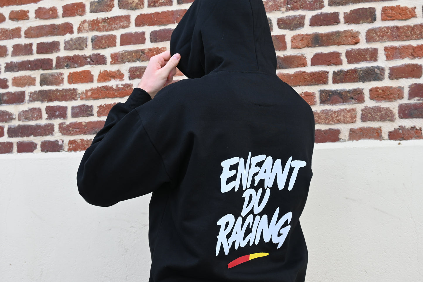 Homme de dos portant un sweat à capuche noir Enfant du Racing avec marquage blanc et détail sang et or, debout devant un mur de briques rouges typique du Nord.