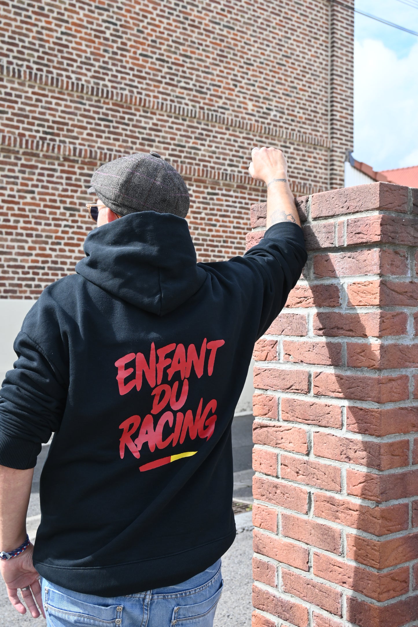 Homme de dos portant un sweat à capuche noir Enfant du Racing avec marquage rouge et détail sang et or, accoudé à un mur de briques dans un décor typique du bassin minier.