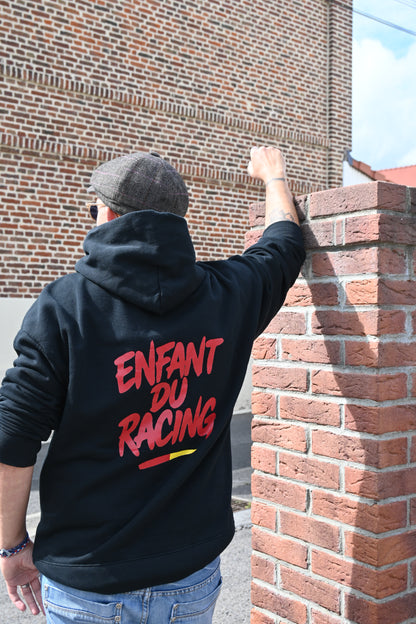 Homme de dos portant un sweat à capuche noir Enfant du Racing avec marquage rouge et détail sang et or, accoudé à un mur de briques dans un décor typique du bassin minier.