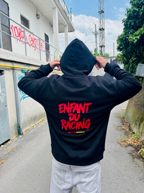 Homme de dos portant un sweat à capuche noir Enfant du Racing, avec impression rouge et détail rouge et or, dans une rue urbaine avec graffitis en arrière-plan.
